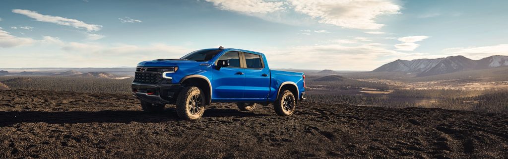 A blue 2025 Chevrolet Silverado 1500 navigating rugged sandy desert terrain near Seminole, OK, with towering mountains in the background and a bright, sunny sky overhead.