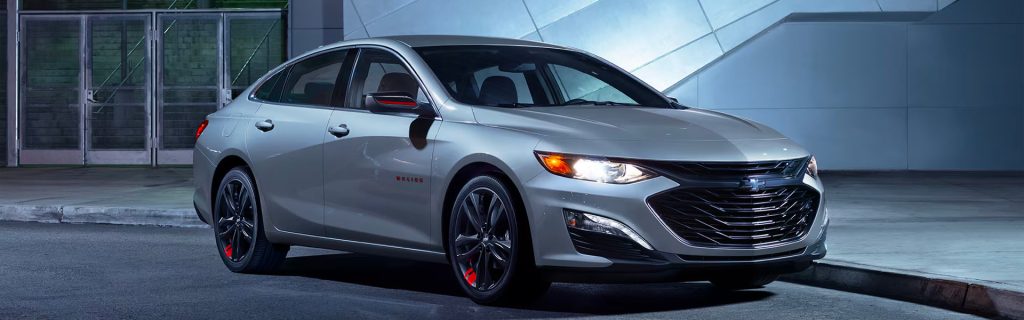 A 2025 silver Chevrolet Malibu with its headlights on, parked along a cement sidewalk outside a closed shopping mall near Seminole, OK. The mall entrance, featuring glass doors and darkened windows, is visible in the background.