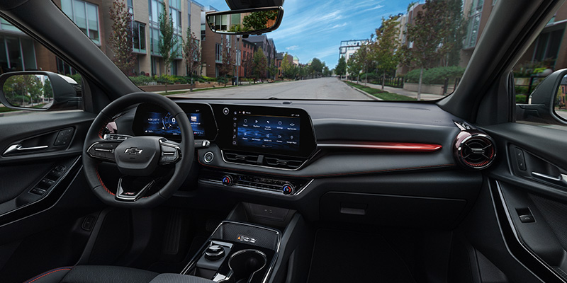A passenger-side view of the 2025 Chevrolet Equinox, parallel parked along a sidewalk on a neighborhood road lined with apartments near Seminole, OK. The interior features a sleek black and gray color scheme with leather seats. The infotainment screen and digital dash display are illuminated, while red ambient lighting adds a sophisticated touch to the passenger-side dashboard.