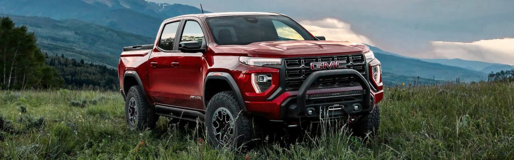 A vibrant red 2025 GMC Canyon parked in a grassy field near Seminole, OK, with towering dark green forest trees on the back left and a scenic mountainside in the background.