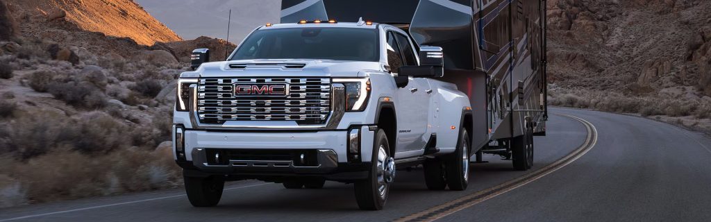 A powerful white GMC Sierra HD dually truck hauls a large fifth-wheel trailer along a winding desert highway near Seminole, OK. The truck's bold chrome grille, LED headlights, and rugged design stand out against the rocky terrain and arid landscape, illuminated by the warm glow of the setting sun.