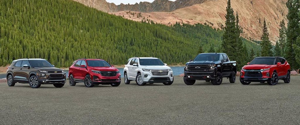 A row of 2025 Chevrolet models, including SUVs and trucks, parked by a serene lakeshore with a breathtaking mountainside in the background. Lush green forests stretch along the mountain near Seminole, OK.