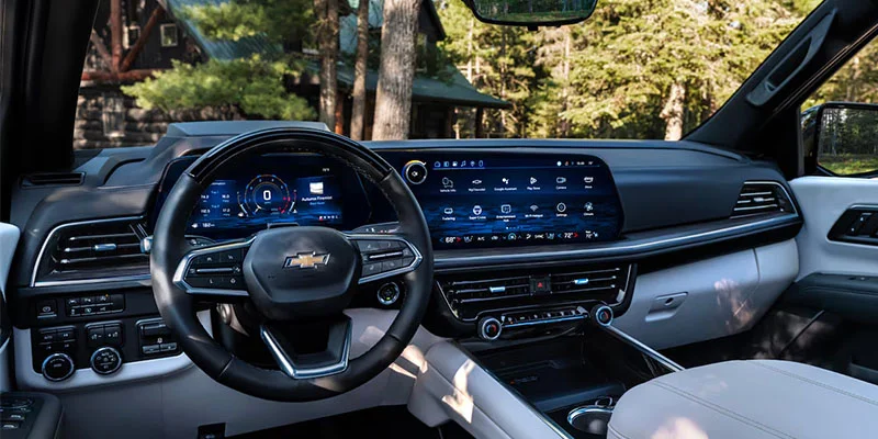Interior of the 2025 Chevrolet Suburban featuring a 17.7-inch infotainment screen, with black and beige seating, parked near tall forest trees in Seminole, OK.