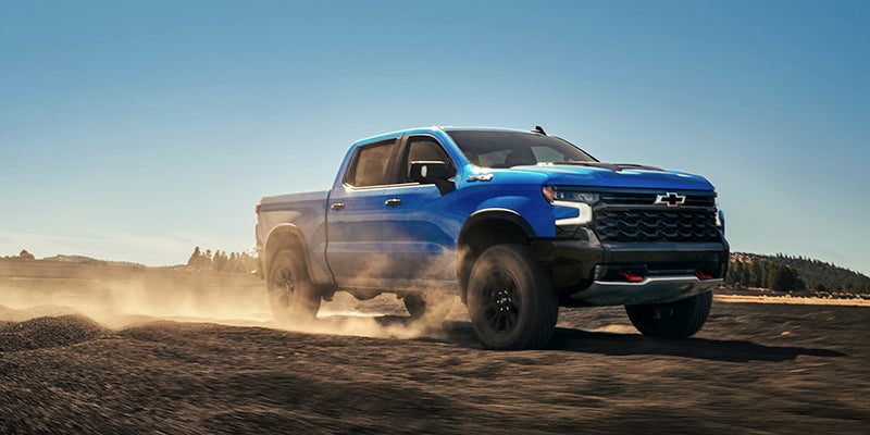 A blue 2025 Chevrolet Silverado 1500 driving fast through black sand and dirt in a desert landscape near Seminole, OK, during daylight.