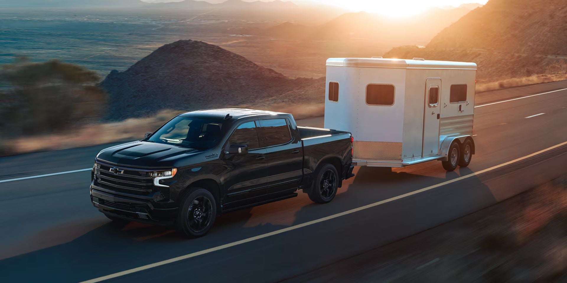 Black 2025 Chevrolet Silverado 1500 towing a horse trailer down a one-lane highway at sunset, with mountainous views in the background near Seminole, OK.